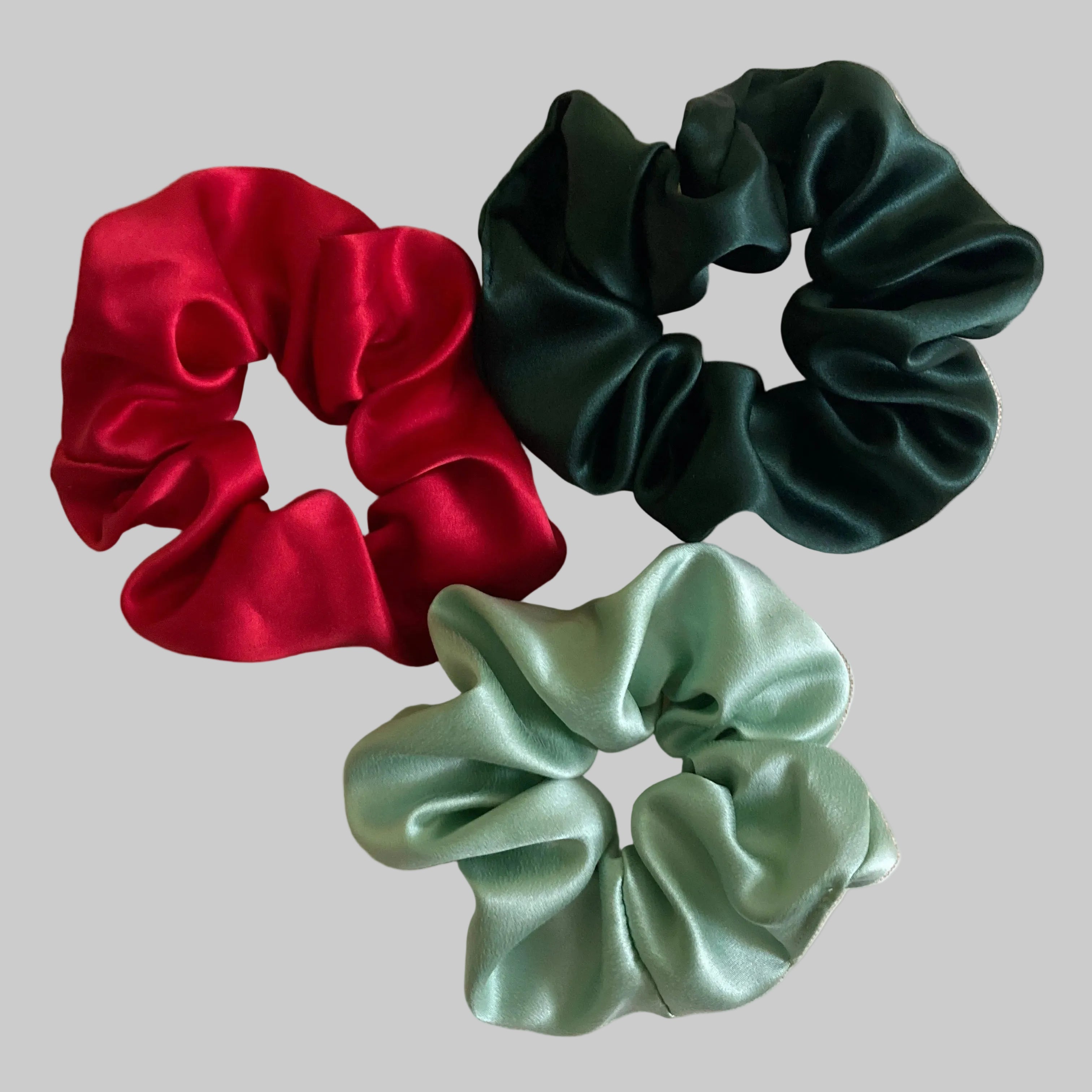 Three satin hair scrunchies in dark green, mint green, and red, arranged on a beige fabric background.